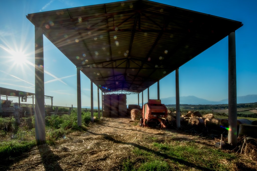 Un capannone agricolo con un trattore rosso e balle di fieno sotto il sole, con vista sui campi e le montagne lontane.