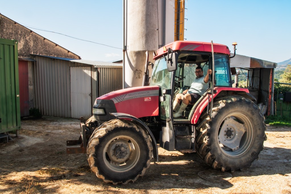 Un trattore rosso parcheggiato in un'area rurale, con un uomo seduto al volante. Sullo sfondo si vedono strutture agricole e un cielo azzurro.