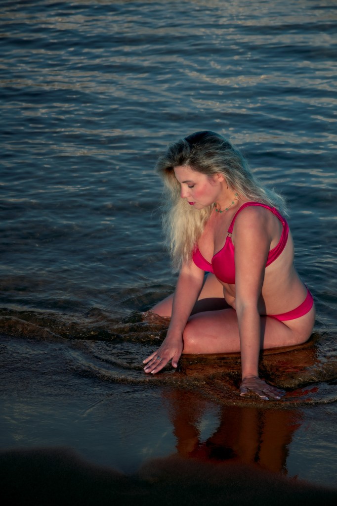 Una donna con capelli biondi seduta in acqua, indossando un bikini rosso, mentre tocca la superficie dell'acqua con una mano.