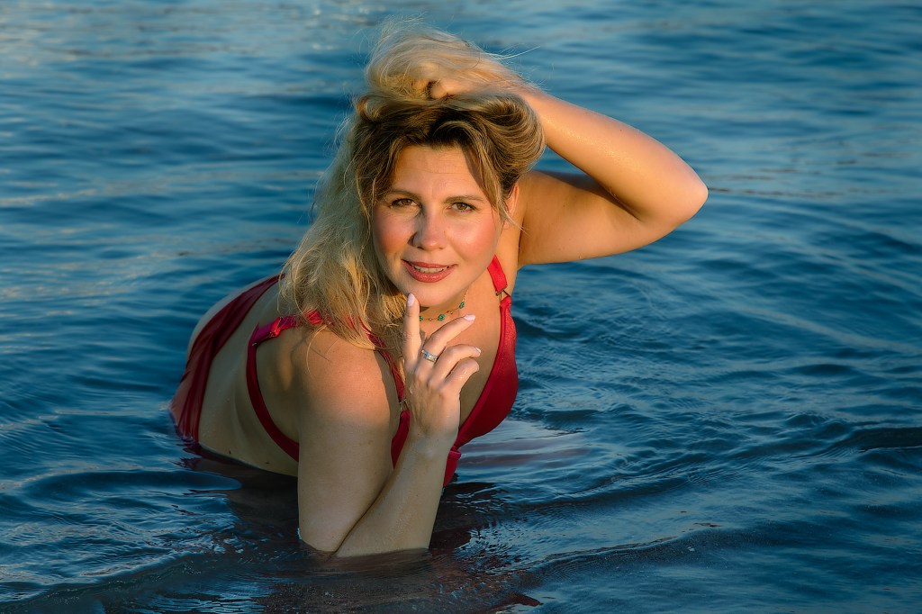 Donna bionda in costume da bagno rosso che posa nell'acqua con un'espressione intrigante.