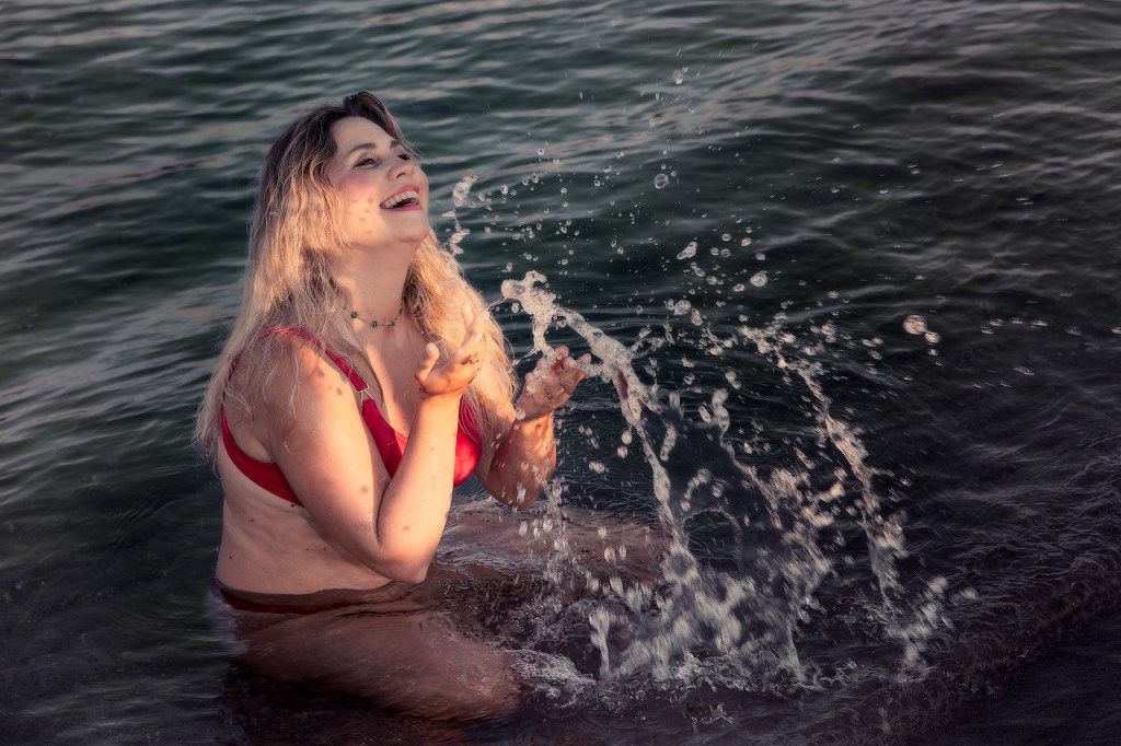 Una donna sorridente in bikini rosso che gioca nell'acqua, spruzzando acqua mentre sta seduta su una roccia.