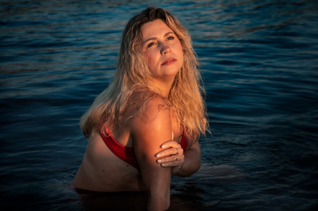 Una donna con capelli lunghi e biondi, vestita con un bikini rosso, si trova nell'acqua blu, guardando con espressione serena verso l'osservatore.