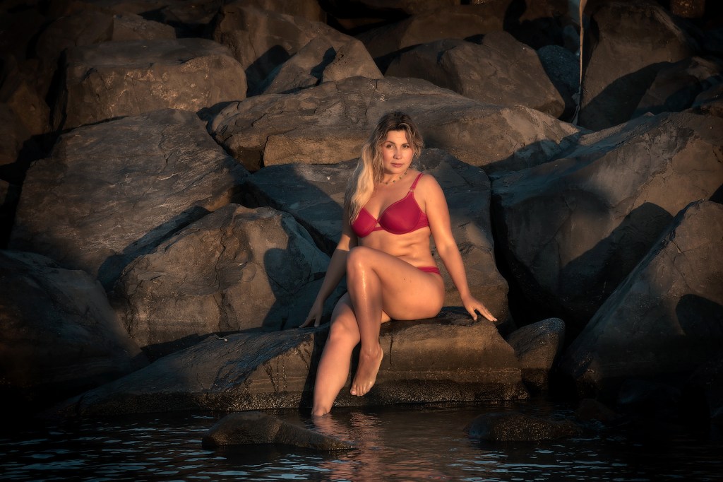Una donna seduta su una roccia bagnata, indossando un reggiseno rosso e mostrando gambe nude, con i capelli mossi, mentre il sole tramonta sullo sfondo.