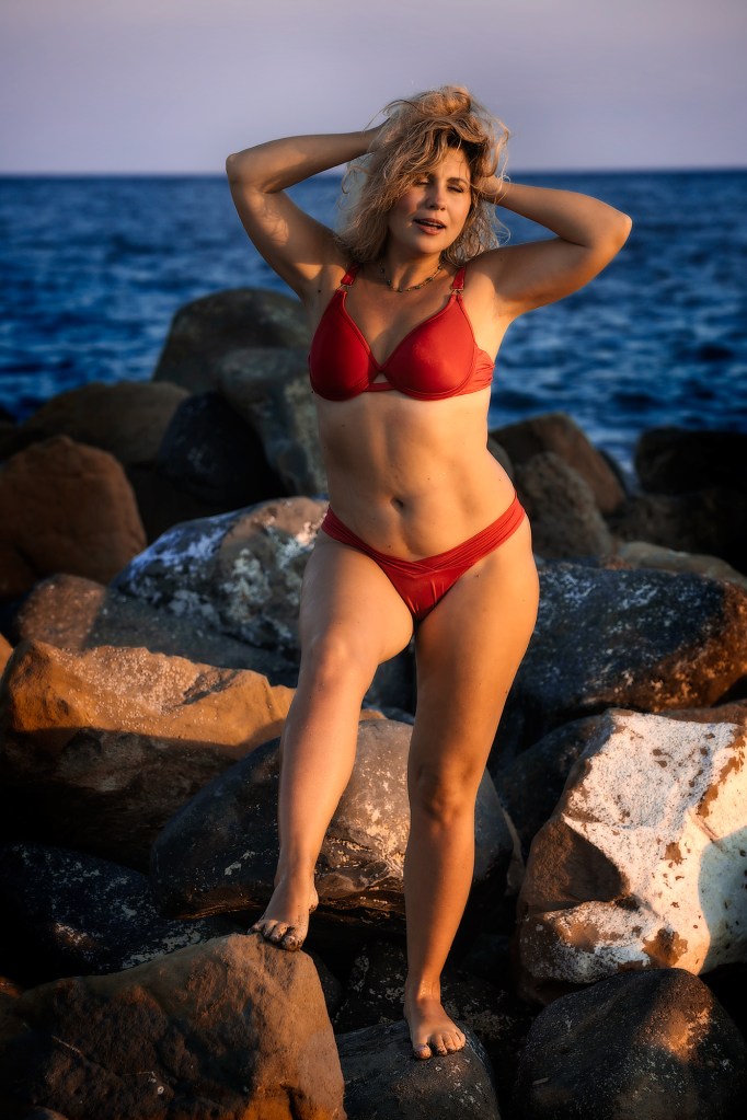 Donna in bikini rosso che posa su rocce vicino al mare, con i capelli mossi e uno sfondo marino al tramonto.