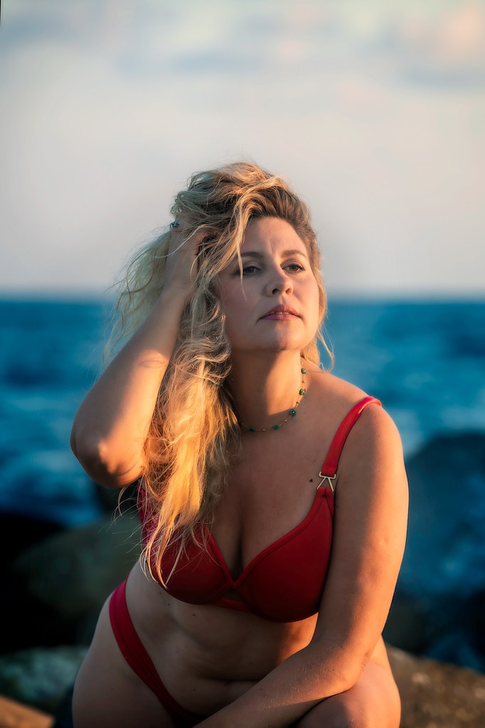 Una donna con capelli biondi mossi indossa un bikini rosso, mentre posa su una roccia con il mare sullo sfondo. La luce del sole illumina il suo viso.
