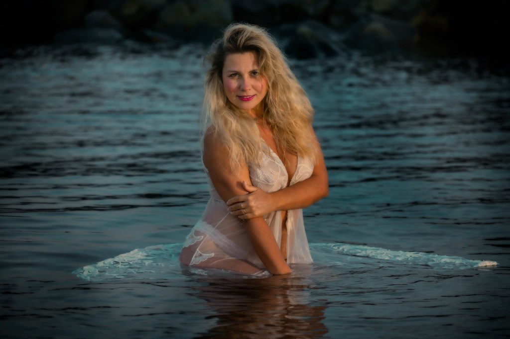 Una donna con lunghi capelli biondi, vestita con un abito trasparente, posa in piedi nell'acqua. Sullo sfondo si vedono delle rocce e la superficie dell'acqua riflette la luce del sole.