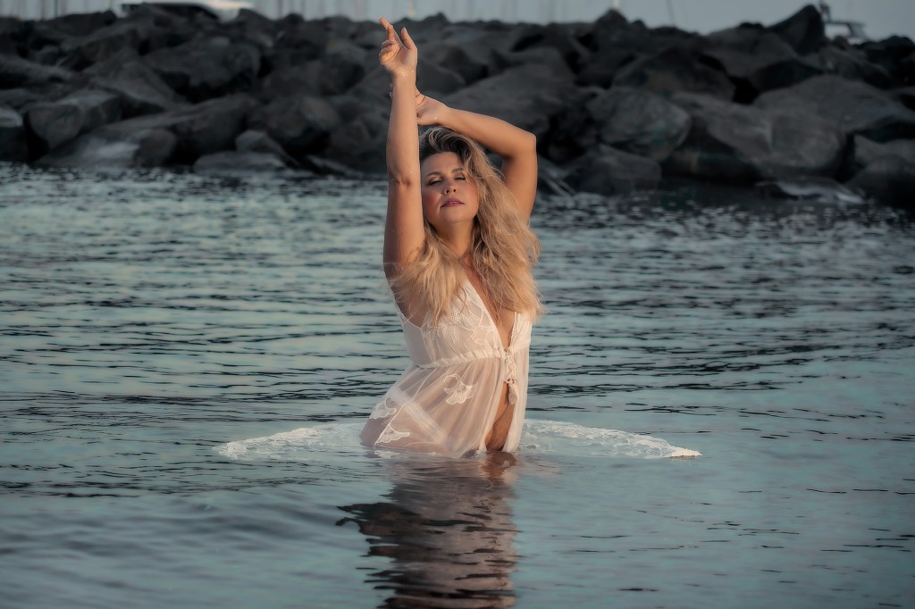 Una donna con capelli lunghi e ricci indossa un vestito bianco trasparente mentre si trova in acqua, sollevando le mani in un gesto dinamico. Sullo sfondo si vedono rocce e una leggera luce naturale.