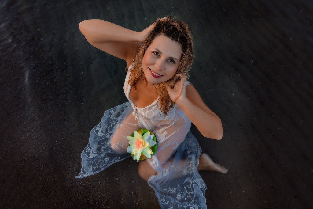 Una donna sorridente seduta nell'acqua con un vestito trasparente e un fiore tra le mani.